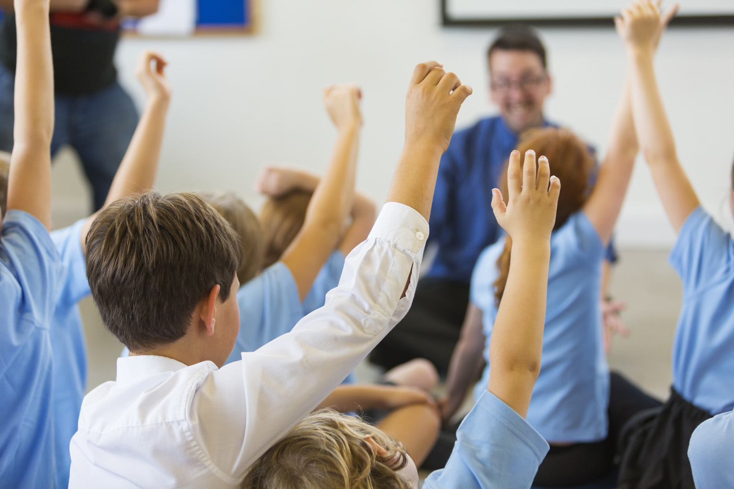 Photo of children with their hands up, eager to answer a question from their teacher
