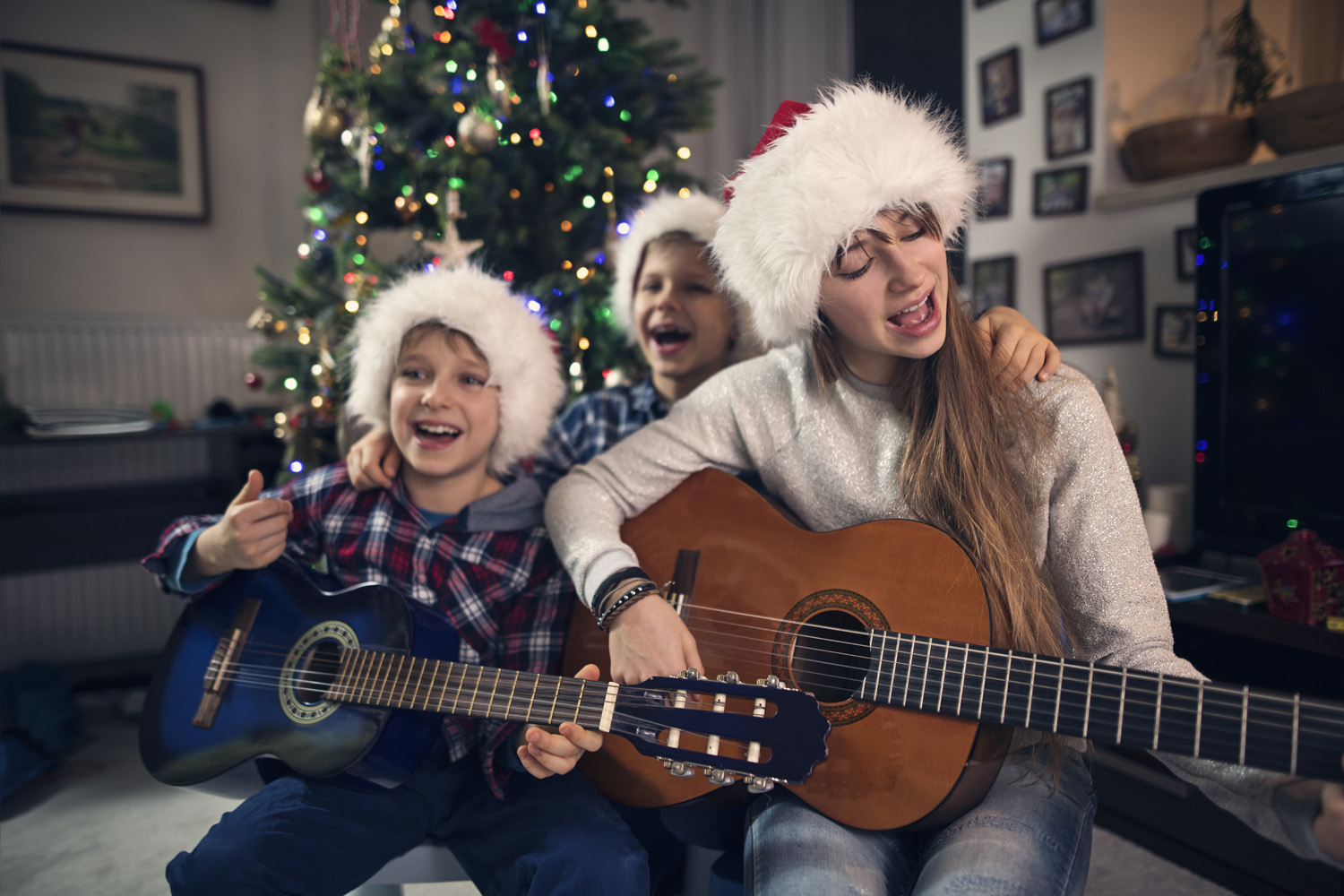 Photo of three children wearing Father Christmas hats sat in front of a Christmas tree playing guitars