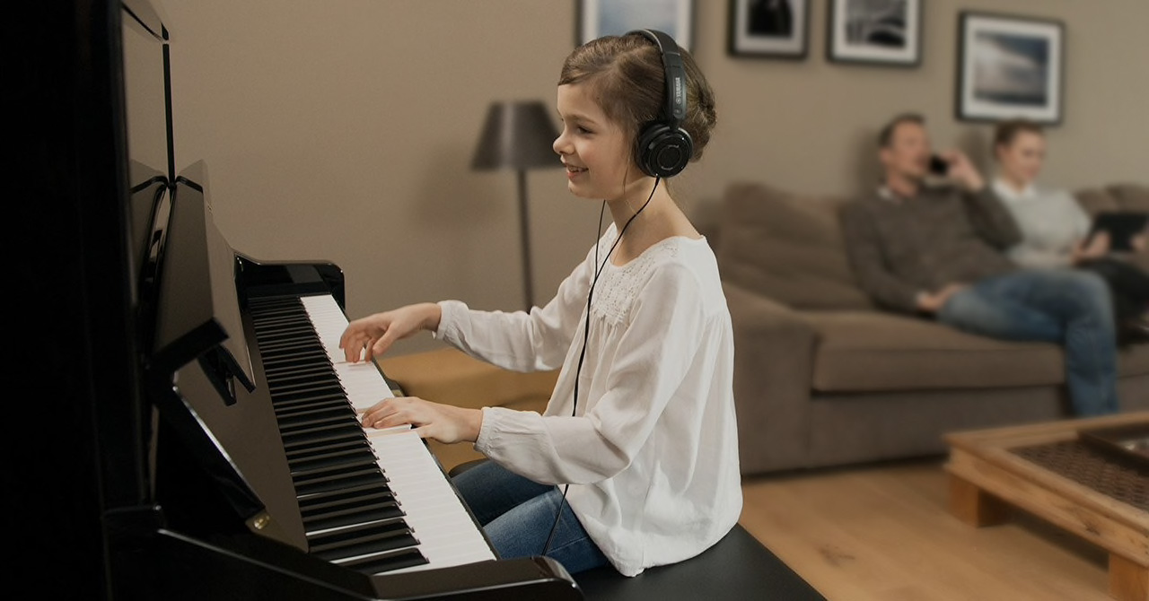 Photo of a girl playing a Silent Piano using headphones whilst her father is on the phone and her mum reads a book on an iPad.